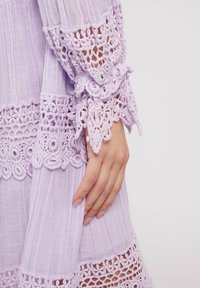 Fliederfarbenes Kleid mit strukturiertem Stoff, filigranem Spitzenbesatz und Rüschensäumen. Nahaufnahme zeigt detaillierte Häkelmuster entlang der Kanten.