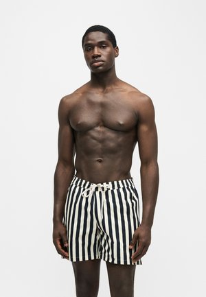 Gespierde, shirtloze man die zwart-witte verticale gestreepte zwemshorts met trekkoord draagt, staand tegen een effen lichte achtergrond.