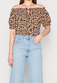 Haut à fleurs décolleté épaule avec fond noir, fleurs orange et roses, manches bouffantes élastiquées ; associé à un jean taille haute bleu clair.