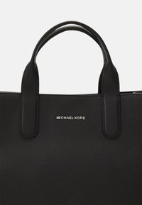 Bolso tote de piel negra con doble asa superior, textura de guijarro y un logotipo metálico de "MICHAEL KORS" en la parte frontal. Diseño limpio y estructurado.