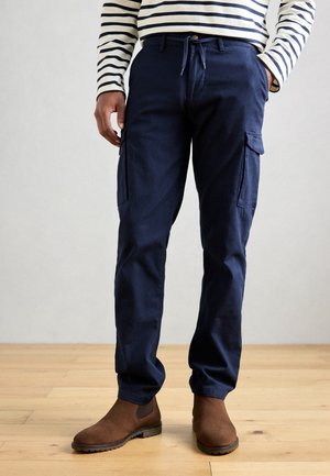 Navy cargo pants made of sturdy fabric, featuring two side pockets, a drawstring waist, and tapered legs. Worn with brown ankle boots.