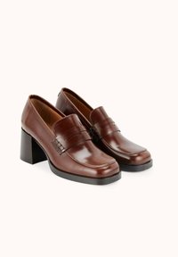 Mocassins à talons en cuir marron avec une texture lisse, un bout carré et une sangle décorative à l'avant. La hauteur du talon est modérée avec une base solide.