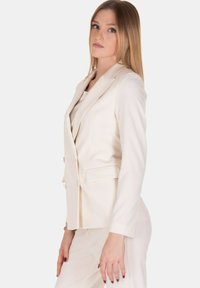 Blazer bianco sartoriale con rever e due tasche frontali. Tessuto liscio e design aderente, abbinato a pantaloni coordinati, per un look coerente.