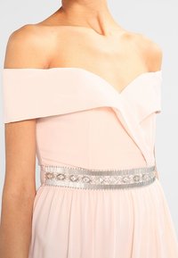 Robe rose clair à épaules dénudées avec un décolleté en V, dotée d'une ceinture ornée d'accent argenté et d'une jupe superposée. Texture de tissu lisse et fluide.