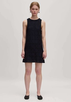 Marineblaues ärmelloses Kleid mit einem strukturierten geometrischen Muster, knielang und mit geradem Saum. Kombiniert mit schwarzen flachen Schuhen.