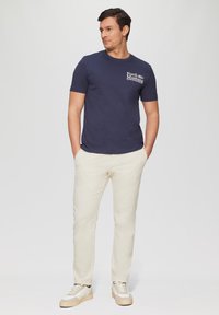 Marineblauwe katoenen t-shirt met "Ford Mustang" graphic op de borst, gecombineerd met crèmekleurige chino's en beige sneakers. Eenvoudige, casual outfit.