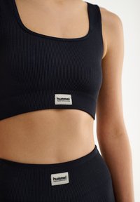 Sostenedor deportivo negro con textura acanalada, de escote cuadrado y una etiqueta del logo de Hummel Performance en la parte inferior. Tejido suave y elástico.