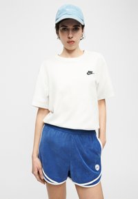 Γυναίκα που φοράει ανοιχτό μπλε καπέλο, λευκό t-shirt της Nike και μπλε αθλητικά σορτς με λευκή επένδυση, σε απλό λευκό φόντο.