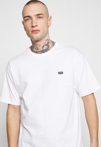 Camiseta blanca de algodón con cuello redondo, mangas cortas y un pequeño parche con el logo negro de Vans en el lado izquierdo del pecho.