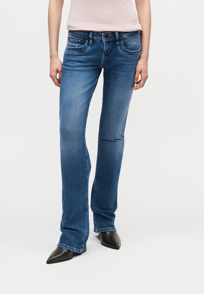 Άτομο που φορά μπλε τζιν καμπάνα και μαύρα παπούτσια με μυτερή μύτη, στέκεται με τα χέρια χαλαρά στα πλάγια μπροστά σε απλό φόντο.