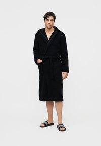 Bărbat purtând un halat de baie negru cu glugă, buzunare și curea, asortat cu șlapi negri Moschino, stând pe un fundal alb simplu.