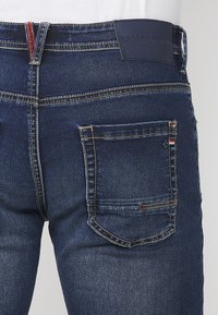 Vue arrière d'un jean en denim bleu foncé avec une poche arrière ornée de détails à rayures rouge, blanc et bleu, ainsi qu'une étiquette de marque en cuir noir.