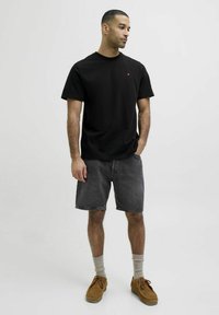 Camiseta de algodón negra con un pequeño logo rojo, combinada con pantalones cortos de mezclilla gris oscuro, calcetines beige y zapatos marrones. Fondo neutro.