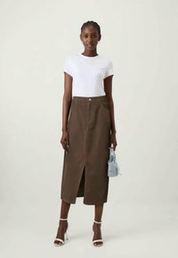 Jupe midi en velours côtelé marron avec fente à l'avant, associée à un t-shirt blanc et des talons blancs à brides, tenant un sac à main bleu clair.