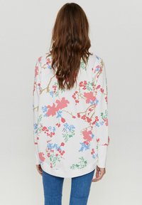 Vit sweatshirt med ett blommönster i rosa, blått och grönt, med långa ärmar och en avslappnad passform. Kombinerad med blå jeans.