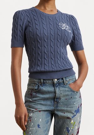 Frau trägt einen kurzärmligen, blau gestrickten Pullover mit weißem Monogramm und blau gesprenkelte Jeans mit Händen in den Taschen.