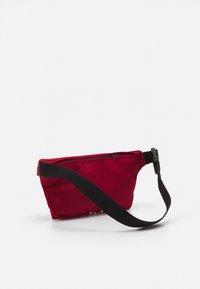 Marsupio in tessuto rosso con una tracolla nera regolabile e una chiusura con zip. Presenta una superficie liscia e una forma compatta, leggermente curva.