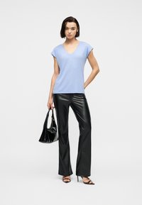 Γυναίκα που φοράει ανοιχτό μπλε μπλουζάκι με V-λαιμόκοψη, μαύρο δερμάτινο παντελόνι, μαύρα ψηλοτάκουνα σανδάλια, κρατώντας μια μαύρη τσάντα, στέκεται μπροστά σε μονόχρωμο φόντο.