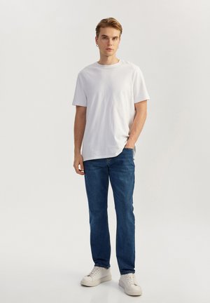 Camiseta blanca de algodón, de manga corta, cuello redondo, combinada con jeans de mezclilla azul, de corte recto, usada con zapatillas blancas, diseño sencillo.