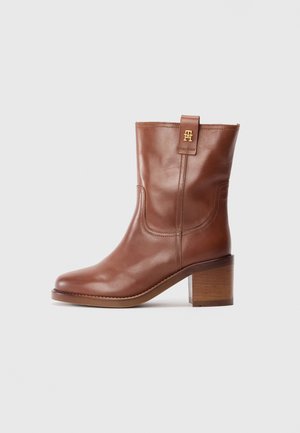 Brown leather mid-calf boot with block heel and gold logo tab on side, shown in profile on a white background.