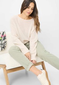 Cremefarvet strikket sweater med lange ærmer, lysegrønne fløjlsbukser og hvide sneakers. Sidder på en struktureret, lysbænk.