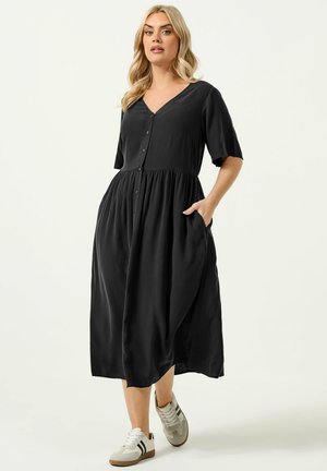 Kvinde med blondt hår iført en sort midi-kjole med knapper foran og hvide sneakers med sorte striber, poserer mod en lys baggrund.