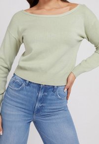 Jasnozielony sweter z odkrytymi ramionami wykonany z gładkiej tkaniny, z długimi rękawami i dopasowaną talią, zestawiony ze średnio wypranymi dżinsami.