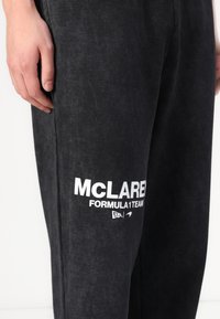 Melni sporta bikses ar mīkstu tekstūru, kuru kreisajā pusē ir balts izdrukāts logo "MCLAREN FORMULA 1 TEAM". Vienkāršs dizains, brīva piegriezuma.