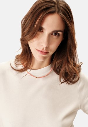 Collier en perles de corail avec des accents dorés porté sur un sweat-shirt crème, présentant un design simple et une texture lisse.