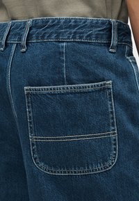 Πίσω τσέπη από σκούρο μπλε τζιν με ορατή ραφή, θηλιές για ζώνη και ένα μέρος από ένα γκρι πουκάμισο που είναι φορεμένο μέσα στην μέση.