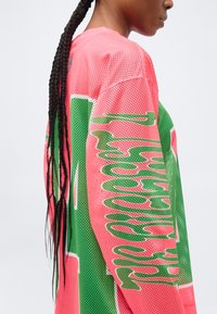 Άτομο με μακριά πλεξούδα που φορά ροζ και πράσινη αθλητική φανέλα από δίχτυ, με μερικώς ορατό κείμενο στο μανίκι και την πλάτη.