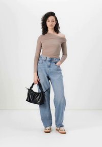 Bež majica dugih rukava s otvorenim ramenima u kombinaciji s svijetloplavim trapericama visokog struka; drži crnu torbu s teksturom; bež cipele s uzorcima.