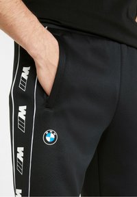 Černé sportovní kalhoty s hladkým povrchem, vybavené bílými bočními pruhy, logy BMW a M, a detaily boční kapsy.