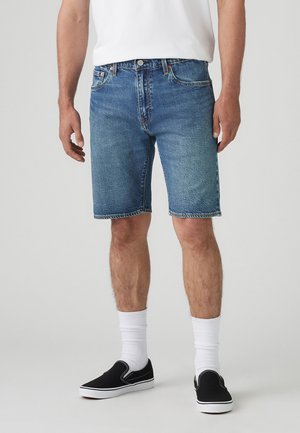 Homme portant un short en jean bleu mi-long, des chaussettes blanches, des chaussures noires sans lacets et un t-shirt blanc sur un fond uni.