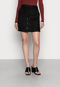 Jupe en daim noir avec fermeture à boutons, taille haute et texture lisse. Assortie avec des mules noires à bout ouvert avec un talon carré.