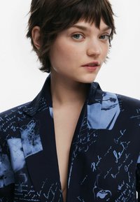 Blazer marine avec des impressions graphiques bleues, présentant des textes et des images manuscrites, design ajusté avec un col en V profond et une texture lisse.