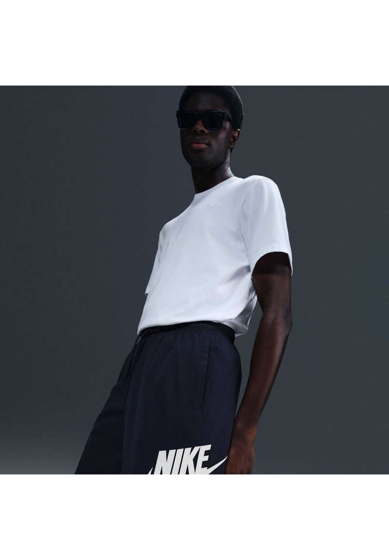 Hombre con gafas de sol negras, camiseta blanca de Nike y pantalones cortos azul marino de Nike, frente a un fondo gris oscuro.