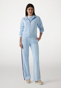 Lichtblauwe zip-up jas met bijpassende broek, gemaakt van gladde stof. De jas heeft een geribde kraag en manchetten, de broek heeft zijstrepen.