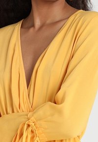 Blusa gialla con scollo a V, maniche lunghe, dettagli arricciati ai polsini e tessuto leggero e fluido, che mette in evidenza la morbidezza e la caduta del materiale.