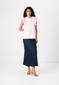 Hellrosa Oberteil mit Rüschensäulen, kombiniert mit einem langen marineblauen Rock. Das Modell trägt goldene Riemchen-Pumps. Glatter Stoffstruktur.