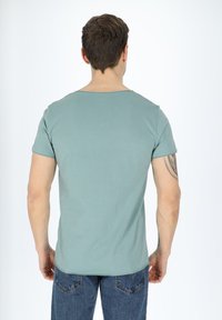 T-shirt di cotone, verde menta, collo rotondo, maniche corte, orlo leggermente curvo, indossata con jeans blu, tatuaggio visibile sul braccio sinistro.