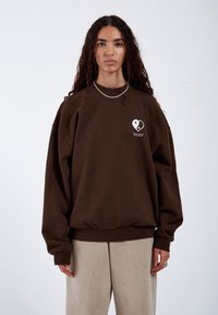 Sweatshirt marrone oversize con scollatura rotonda, caratterizzato da una grafica a cuore bianco sul petto. Tessuto morbido e polsini a costine.