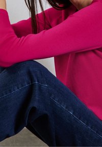 Pitkähihaisen fuksiansävyisen ribbineulospaidassa on rypytetty pinta, ja se on yhdistetty tummansinisiin farkkuihin. Housujen lahkeen malli on väljä, mutta kapenee hieman nilkkaa kohti.