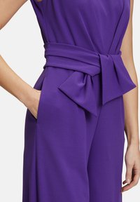 Lila overál, övvel ellátott derékkal és nagy masnival, texturált anyagból, oldalzsebekkel. Letisztult vonalak és szabott fazon.