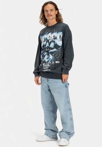 Sudadera gráfica negra con un gran estampado frontal de una calavera y un diseño de relámpagos; combinada con vaqueros sueltos azul claro y zapatillas blancas.