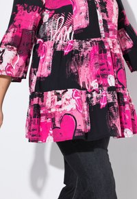 Sort top med pink grafiske mønstre, herunder hjerter og tekst. Har en flagrende design og trekvartærmer. Sammen med mørke jeans.