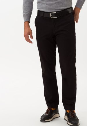 Schwarze Hose mit Gürtelschlaufen und einer silbernen Schnalle, kombiniert mit schwarzen Sneakers mit orangenen Schnürsenkeln. Glattes Material, schlankes Design.