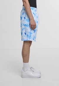 Blå og hvite batikkmønstrede shorts med elastisk midjebånd og snor, vist mot en nøytral bakgrunn. Brukt med hvite joggesko.