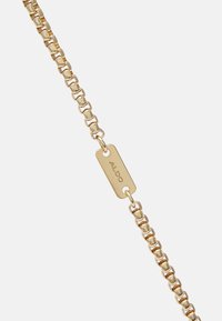 Bransoletka łańcuszkowa w złotym odcieniu z prostokątną zawieszką z wygrawerowanym napisem "ALDO", złożona z przeplatających się kwadratowych ogniw na białym tle.