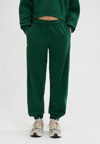 Pantalones de chándal verdes con cinturilla elástica y bolsillos laterales, hechos de material suave. Los puños ajustados en los tobillos completan el diseño.